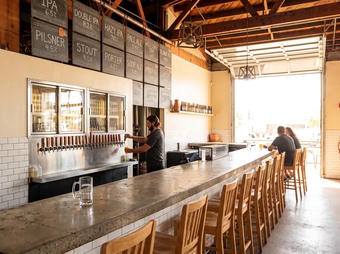Interior of a working brewery taproom, a bartender pulling a pint at a stainless tap wall with customers along the bar.