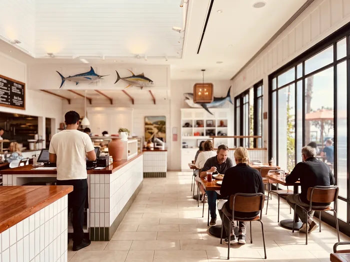 Interior of an airy coastal seafood restaurant, diners at wood tables with mounted game fish on the white shiplap wall.